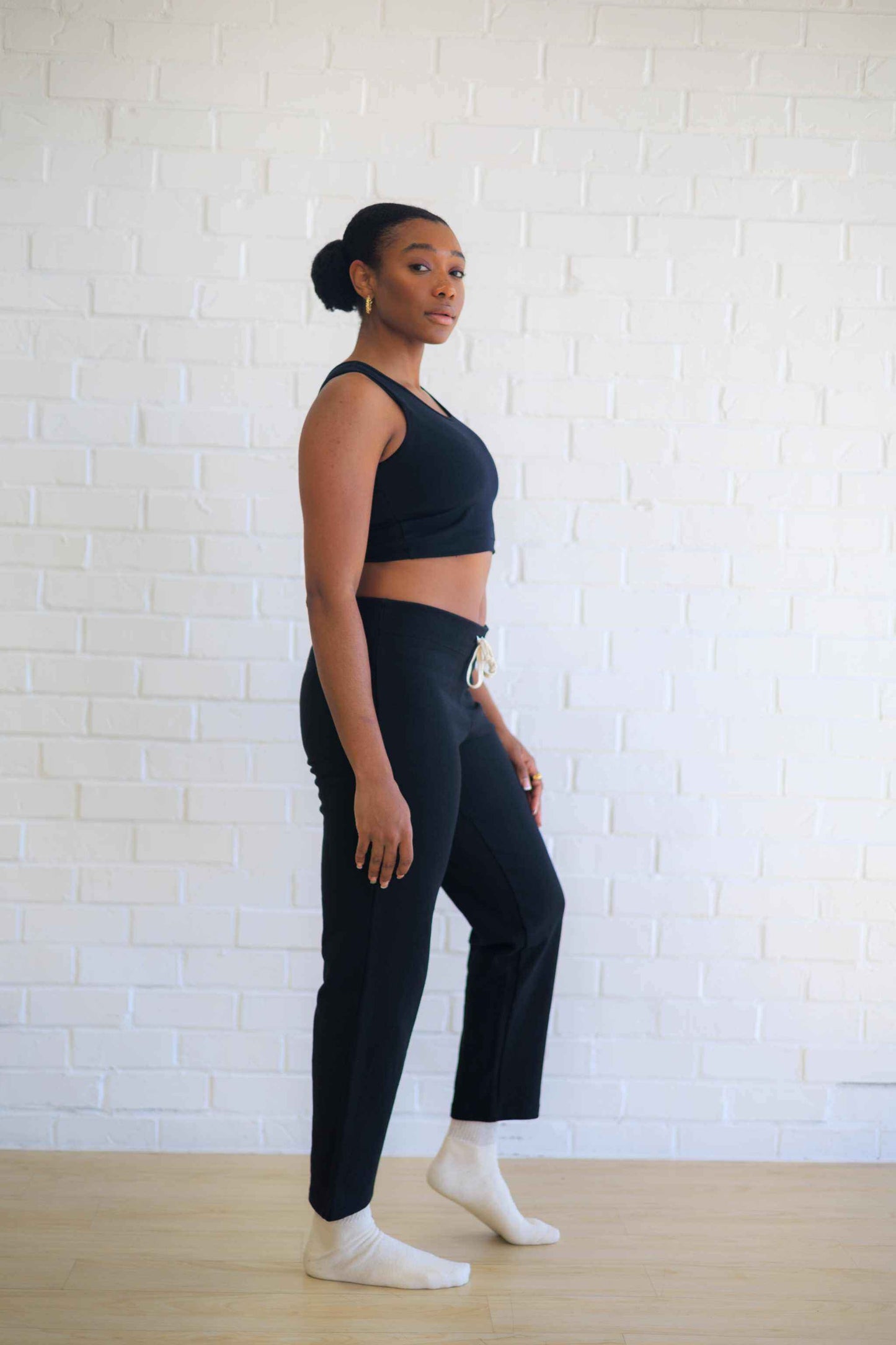 Woman wearing black hemp bralette and black pants standing against white brick wall