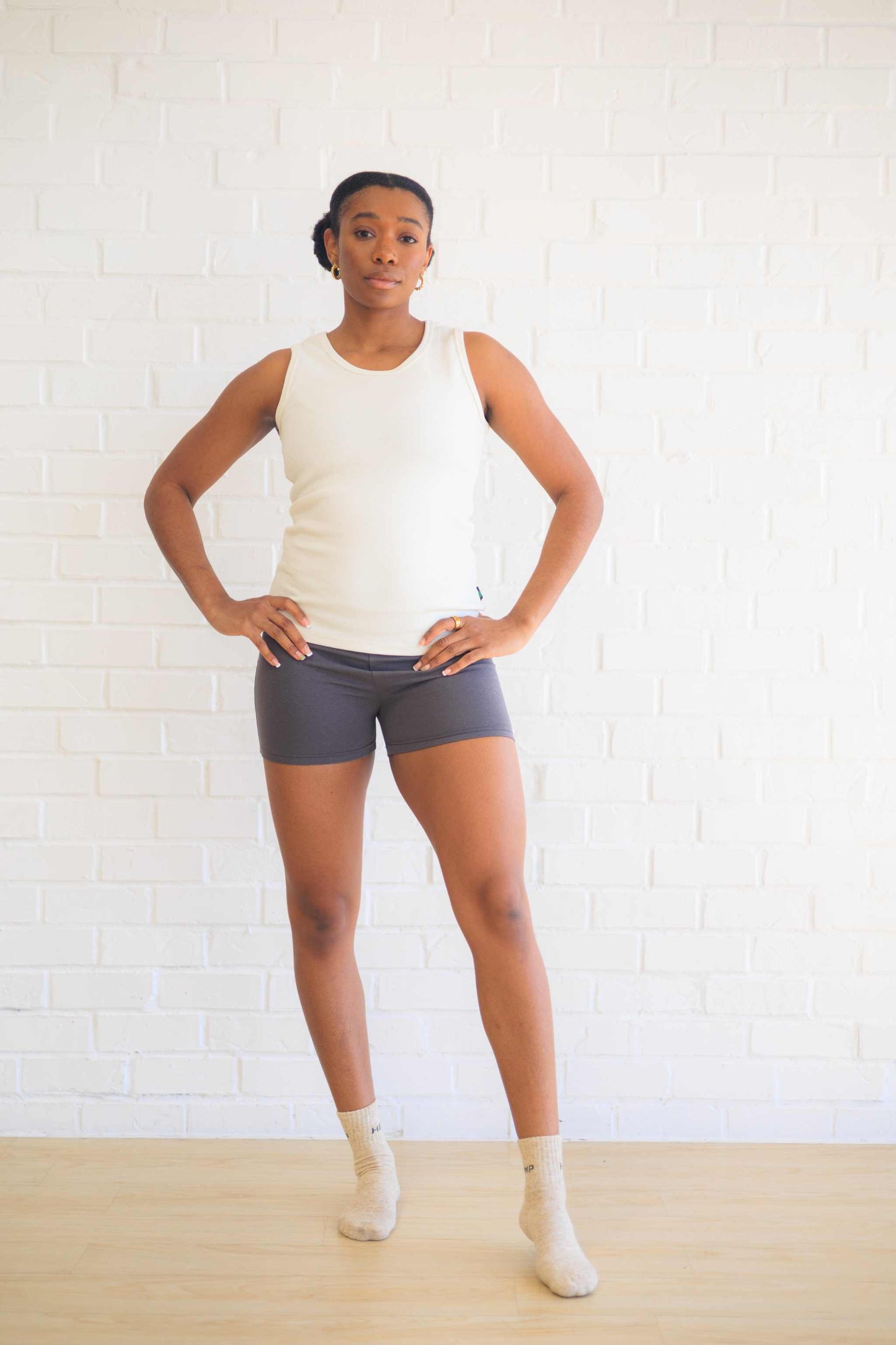 Woman wearing a white cotton tank top with shorts, standing against a white brick wall.