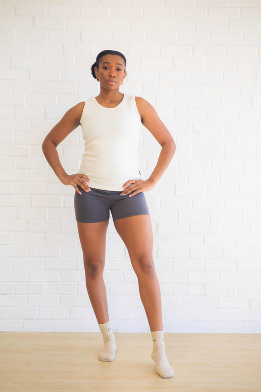 Woman wearing a white cotton tank top with shorts, standing against a white brick wall.