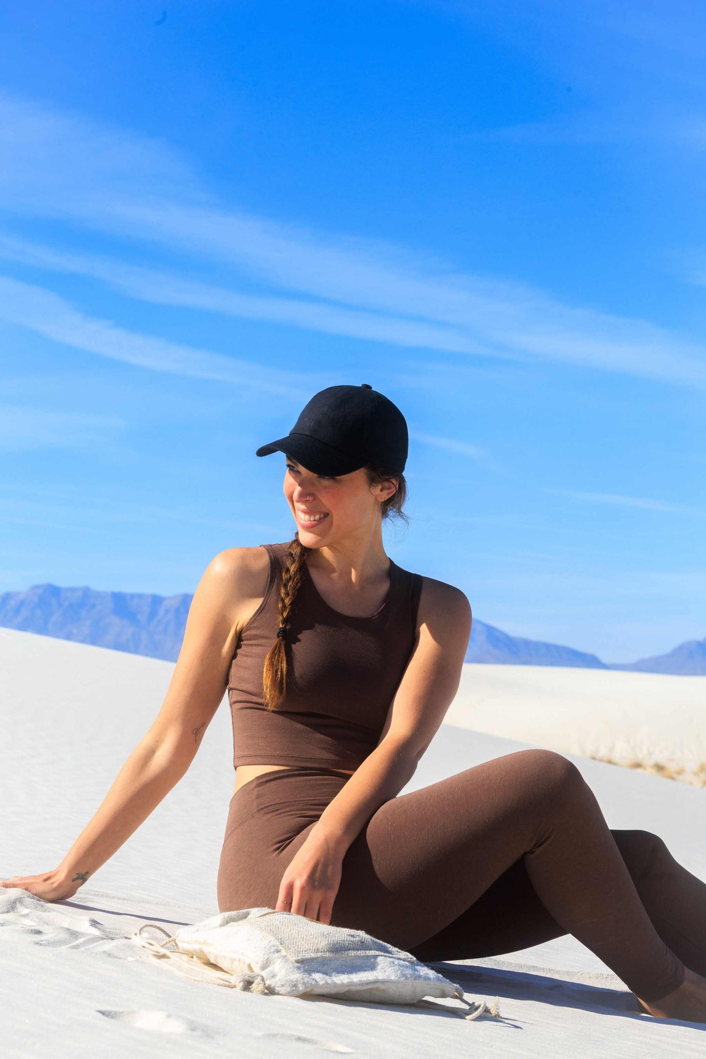 Woman wearing brown hemp crop tank and leggings sitting on sand, background of blue sky and mountains.