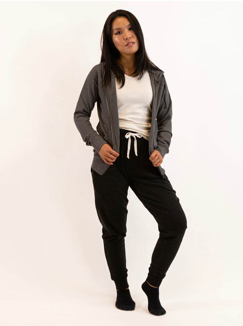 Woman wearing a gray sustainable hemp and organic cotton zip-up hoodie with black joggers and white tank top.