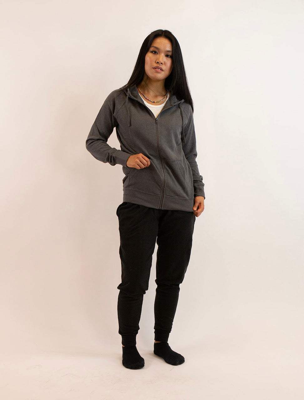 Woman wearing a gray sustainable hemp and organic cotton zip-up hoodie with black pants on a plain background