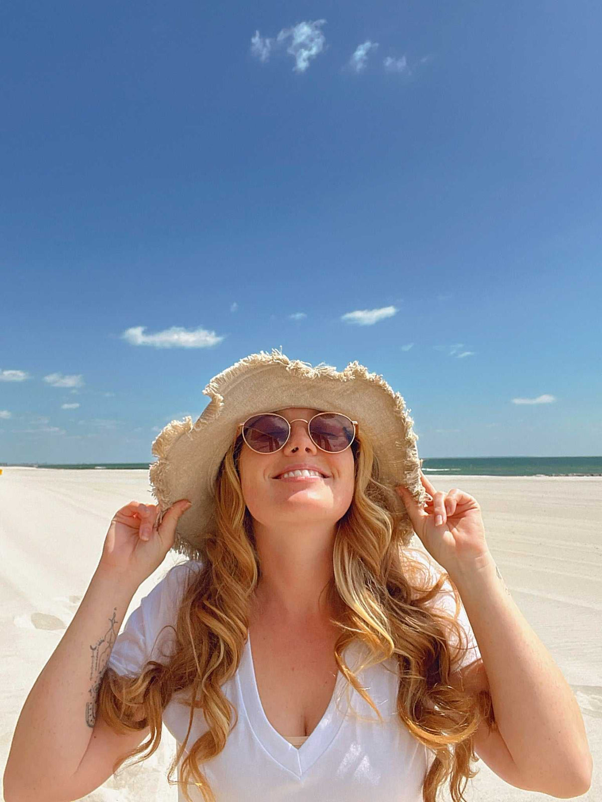 Woman wearing a sun hat and sunglasses on a beach with a clear blue sky.