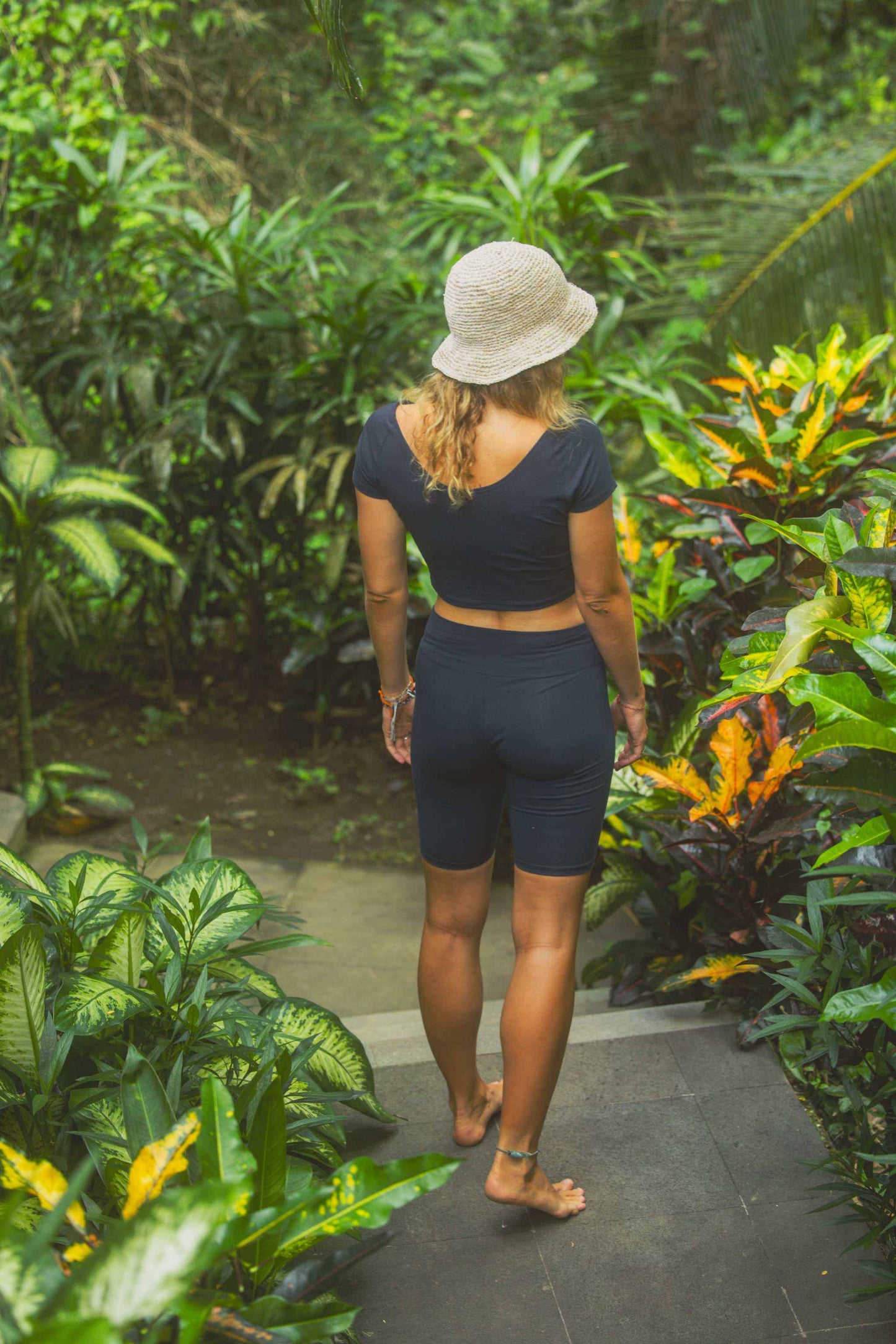 Person walking through a lush tropical garden wearing a black outfit and hat.