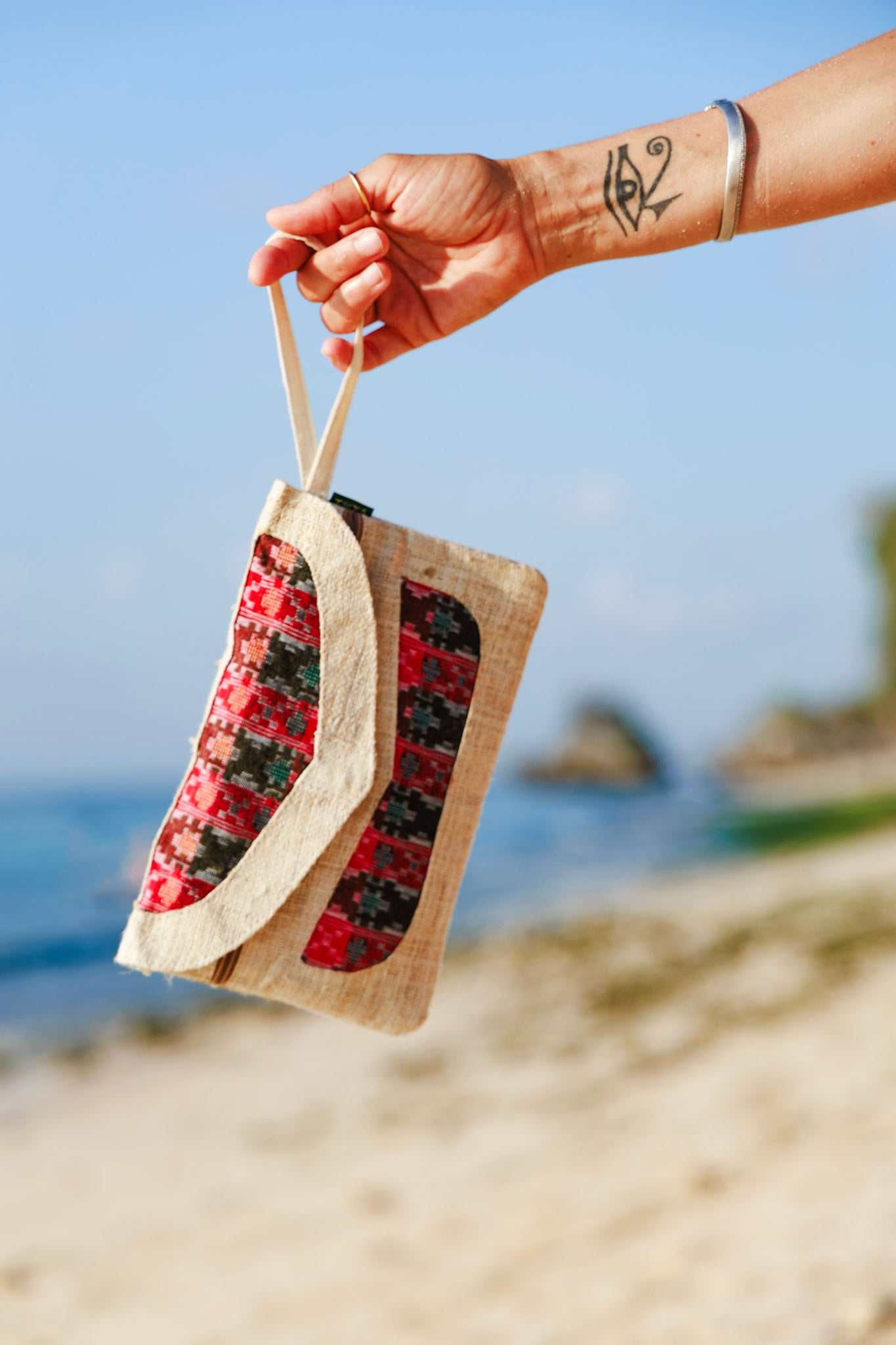 Handmade Himalayan hemp clutch purse held by a hand, featuring red and black patterns, with a hand strap, against a beach background.