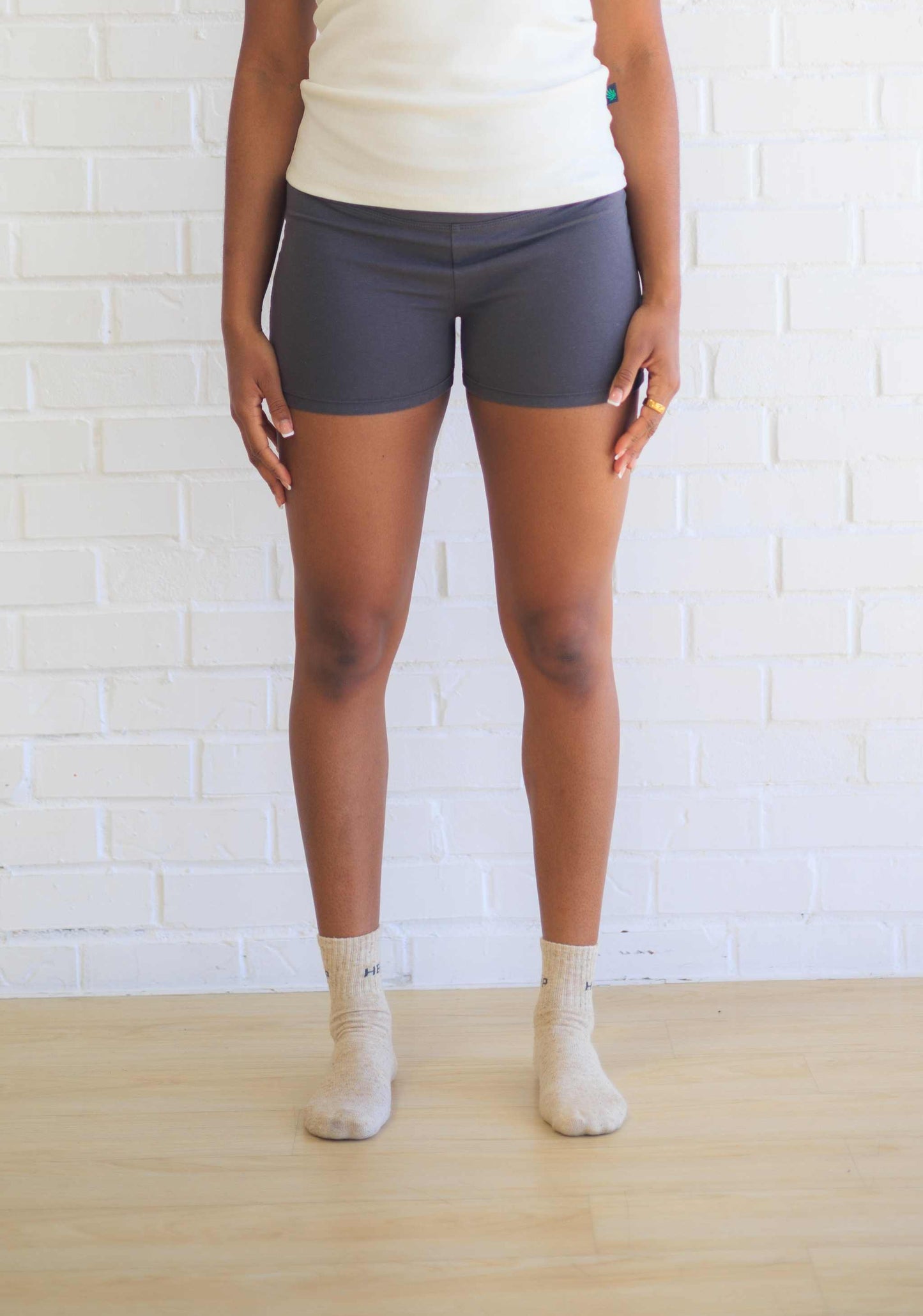 A person standing in front of a white brick wall wearing black active shorts with a visible logo on the waistband, paired with a white top and beige socks.