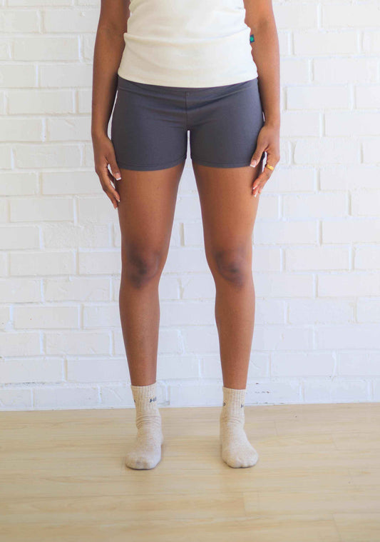 A person standing in front of a white brick wall wearing black active shorts with a visible logo on the waistband, paired with a white top and beige socks.