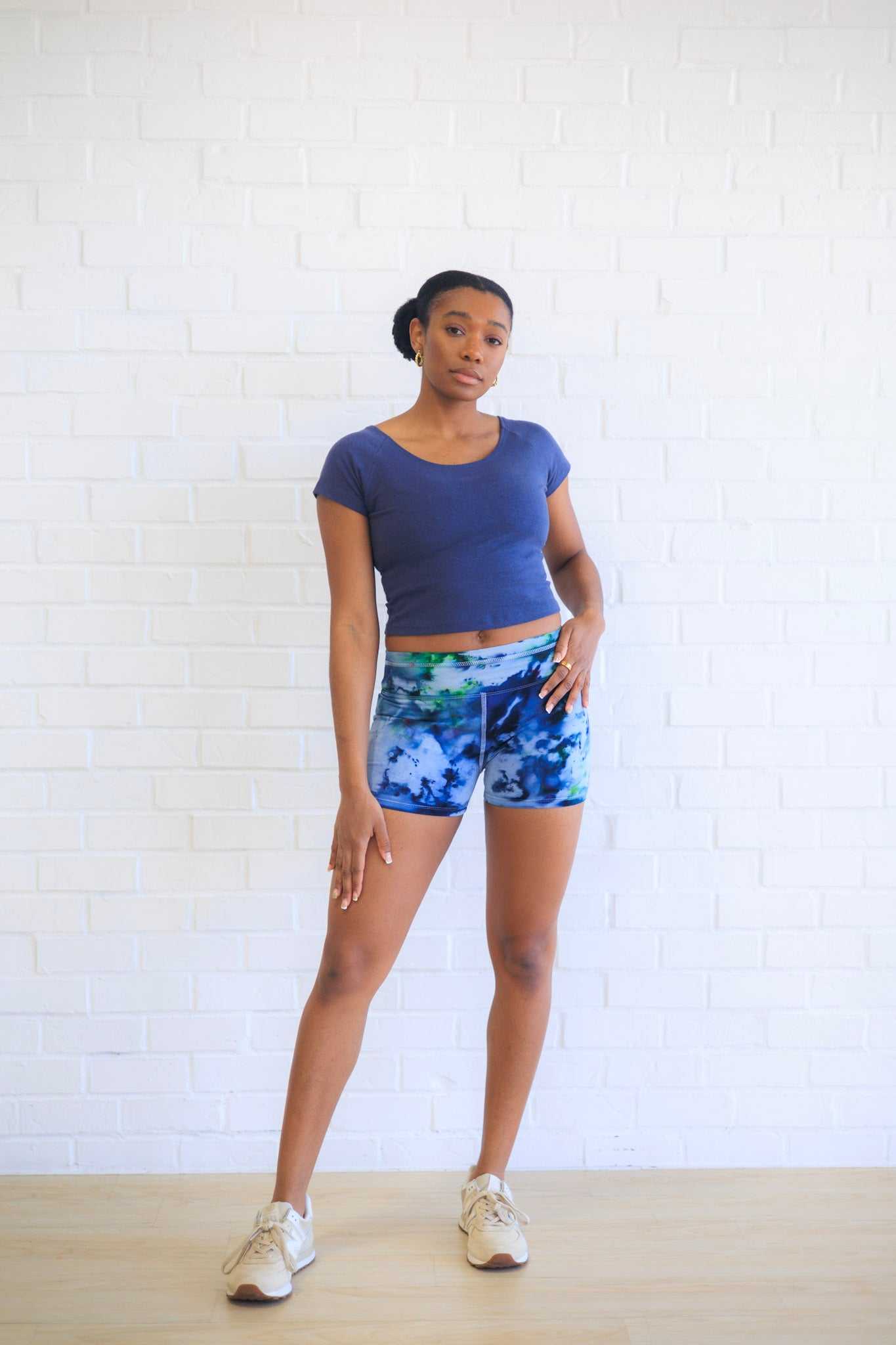 Woman wearing a blue crop top and tie-dye shorts standing against a white brick wall.