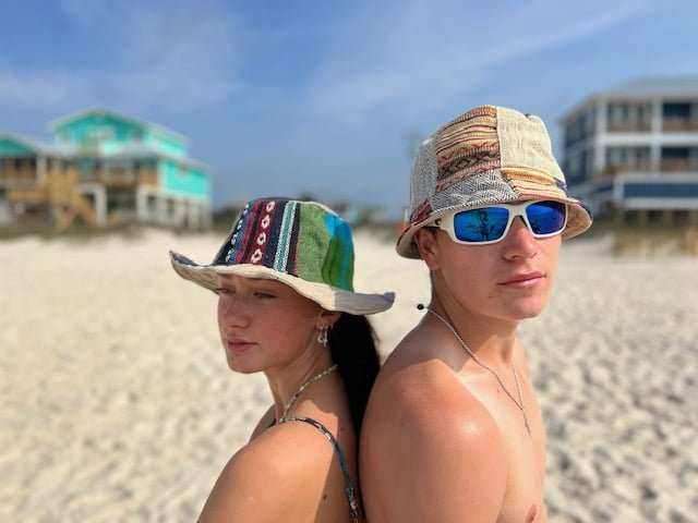 Two people wearing colorful handmade hemp patchwork bucket hats on a beach with buildings in the background