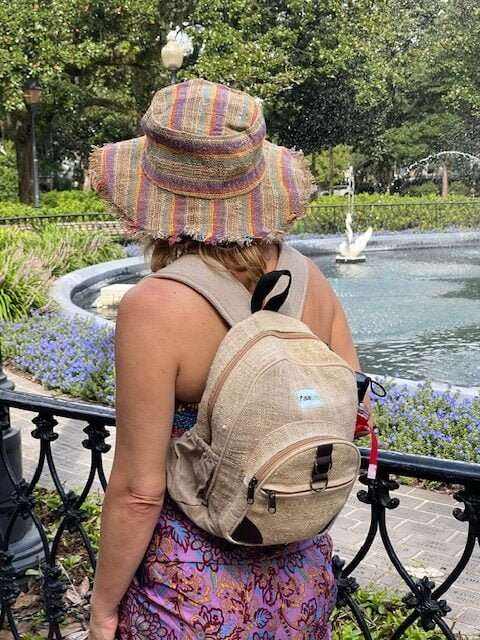 Woman wearing eco-friendly hemp mini-backpack outdoors near fountain with lush greenery and flowers