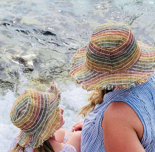 Two people wearing wide brim multi-stripe hemp sun hats by the water, showcasing sustainable sun hats for beach and travel.
