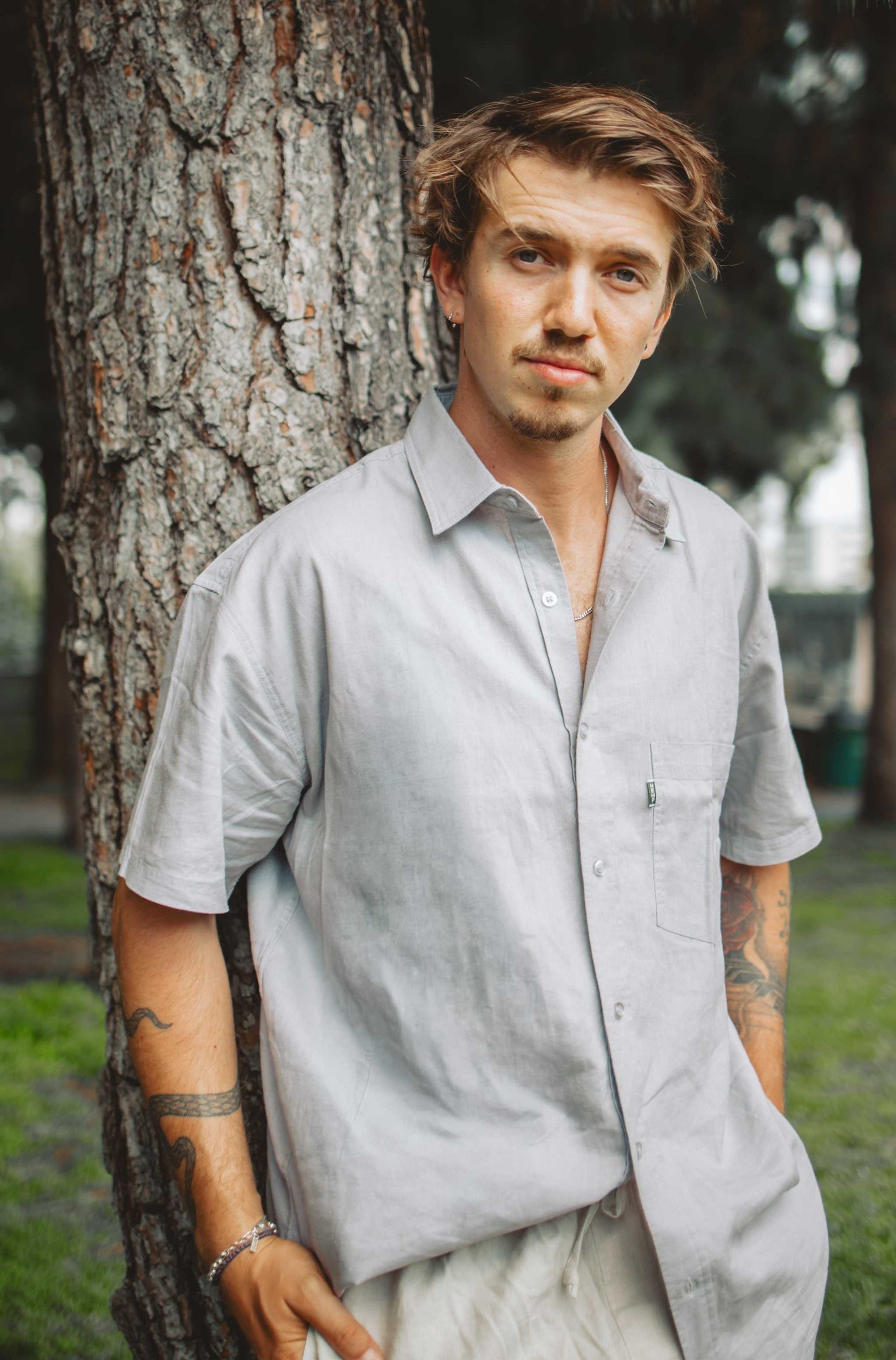 A man with light brown hair and tattoos leans against a large tree trunk outdoors, wearing a light gray short-sleeve linen button-up shirt and light beige trousers.