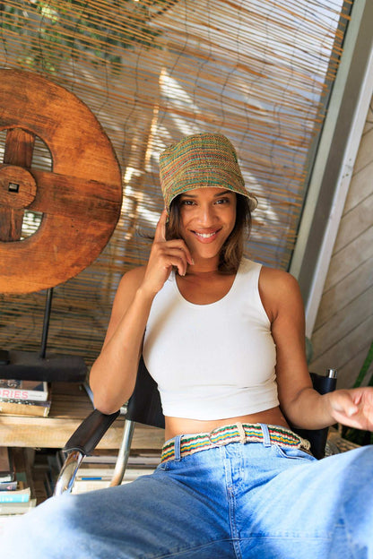 Woman sitting outdoors wearing a white tank top and blue jeans, with a woven wall and wooden table in the background.