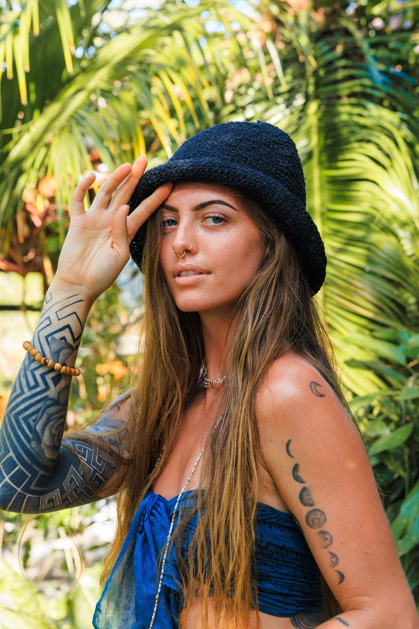 Woman with tattoos wearing a blue top and black hat in front of green foliage