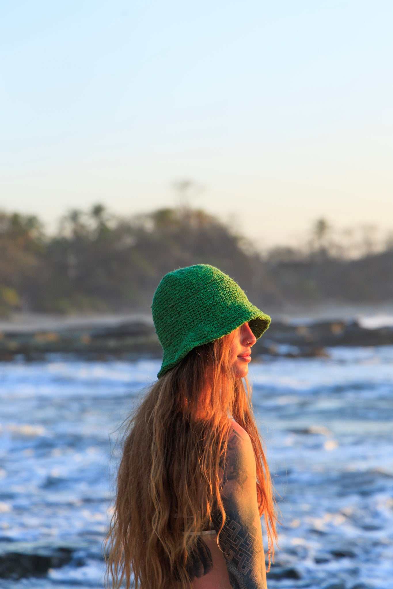 Person wearing a green hat on a beach with ocean waves and trees in the background