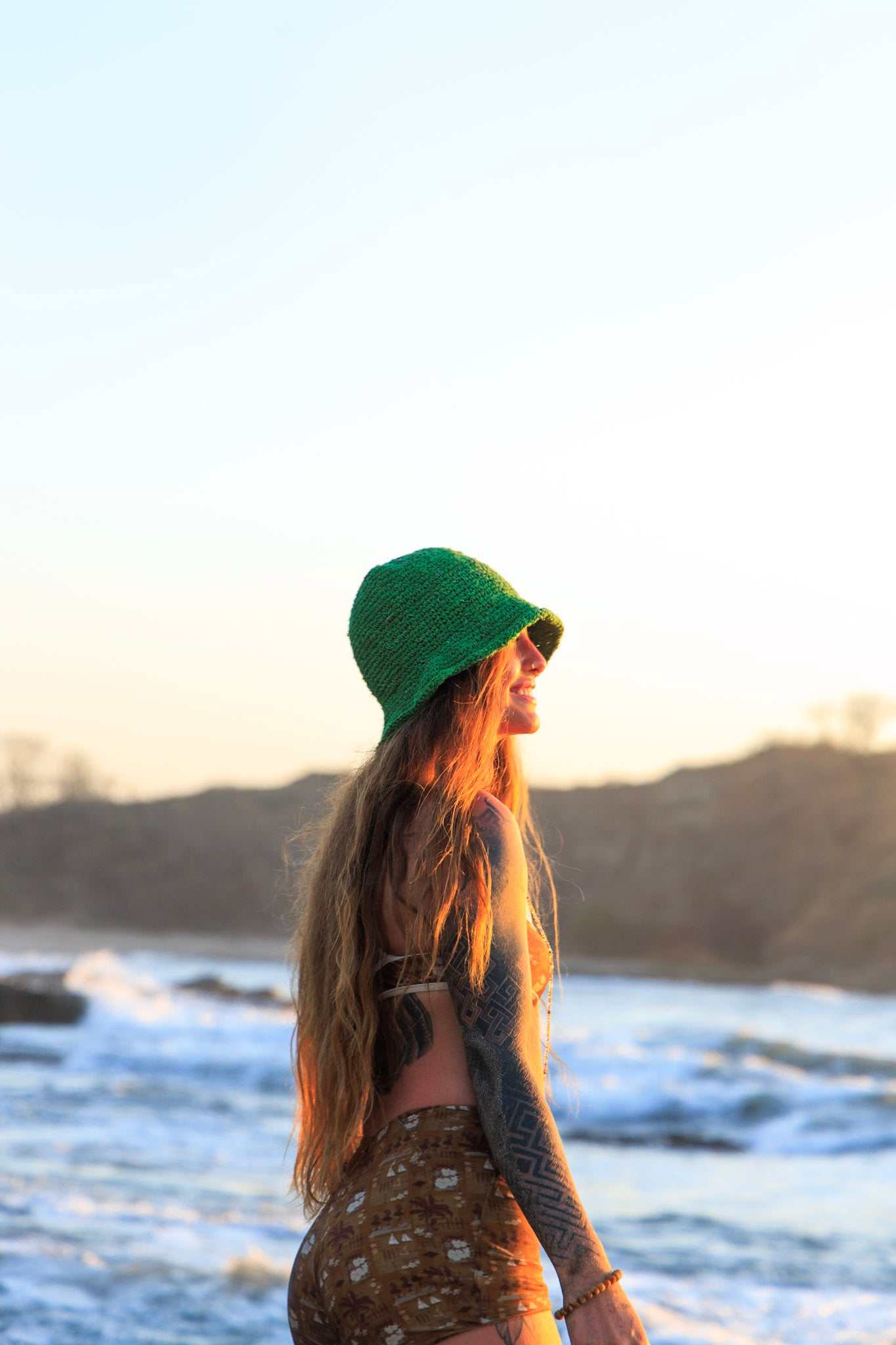 Woman wearing a green hat and patterned dress standing on a beach with ocean waves in the background.
