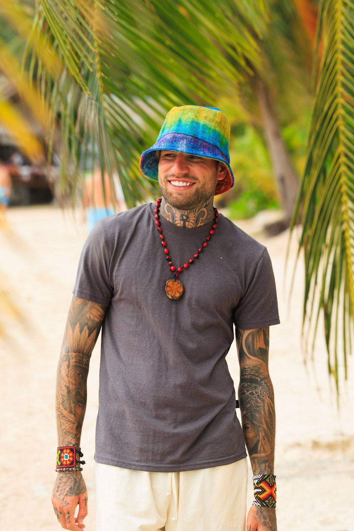 Man wearing a colorful bucket hat and tattoos standing on a beach with palm trees in the background