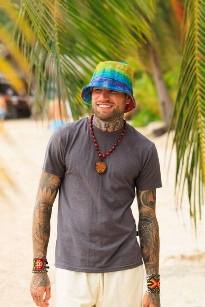 Man wearing a colorful bucket hat and tattoos standing on a beach with palm trees in the background