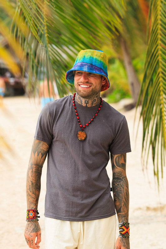 Man wearing a colorful bucket hat and tattoos standing on a beach with palm trees in the background