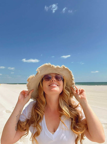 Woman wearing a sun hat and sunglasses on a beach with a clear blue sky.