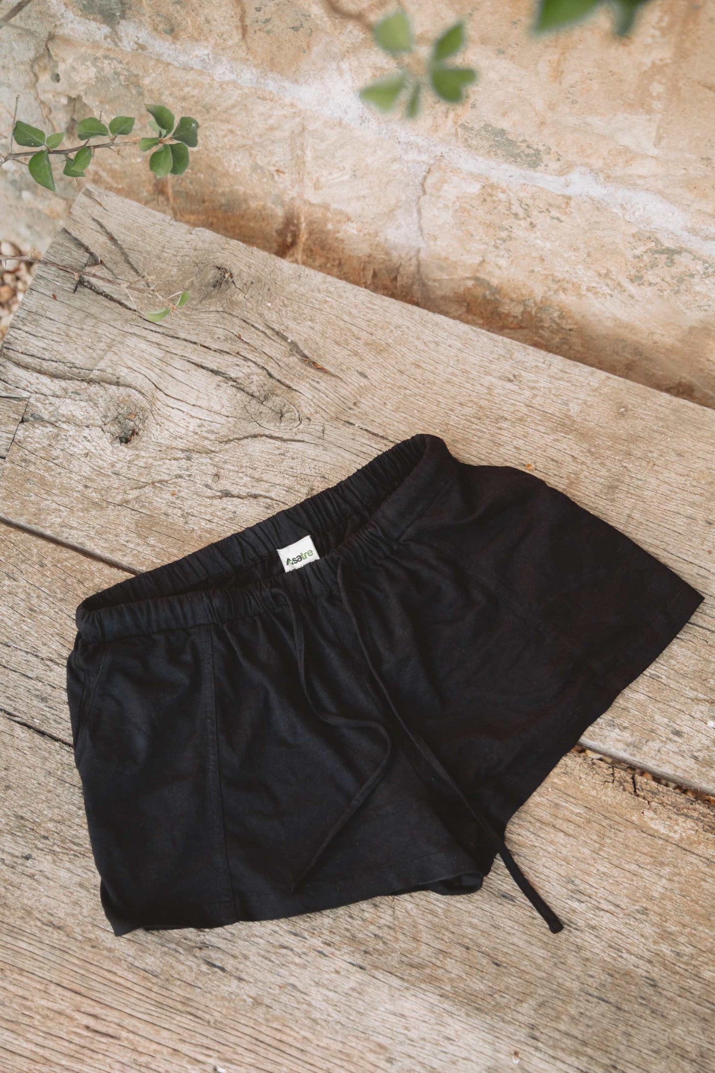 Black drawstring shorts laid flat on a rustic wooden surface with a stone wall and small green leaves in the background.
