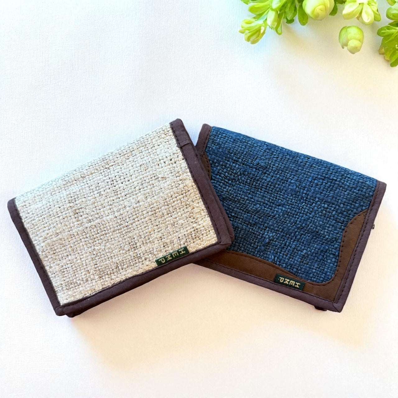 Two fabric wallets, one beige and brown, the other blue and brown, on a light surface with greenery.