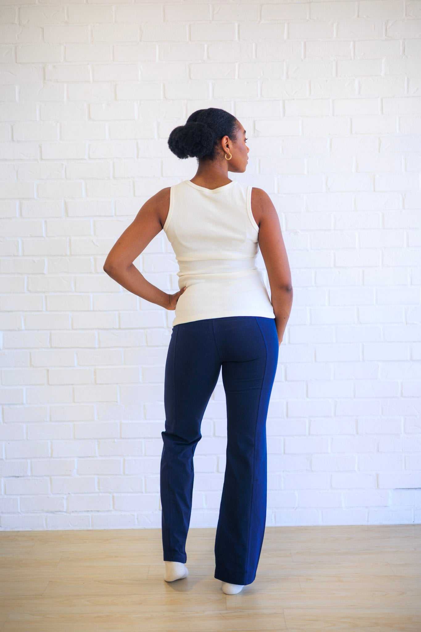 Person wearing a white sleeveless top and blue pants standing against a white brick wall.