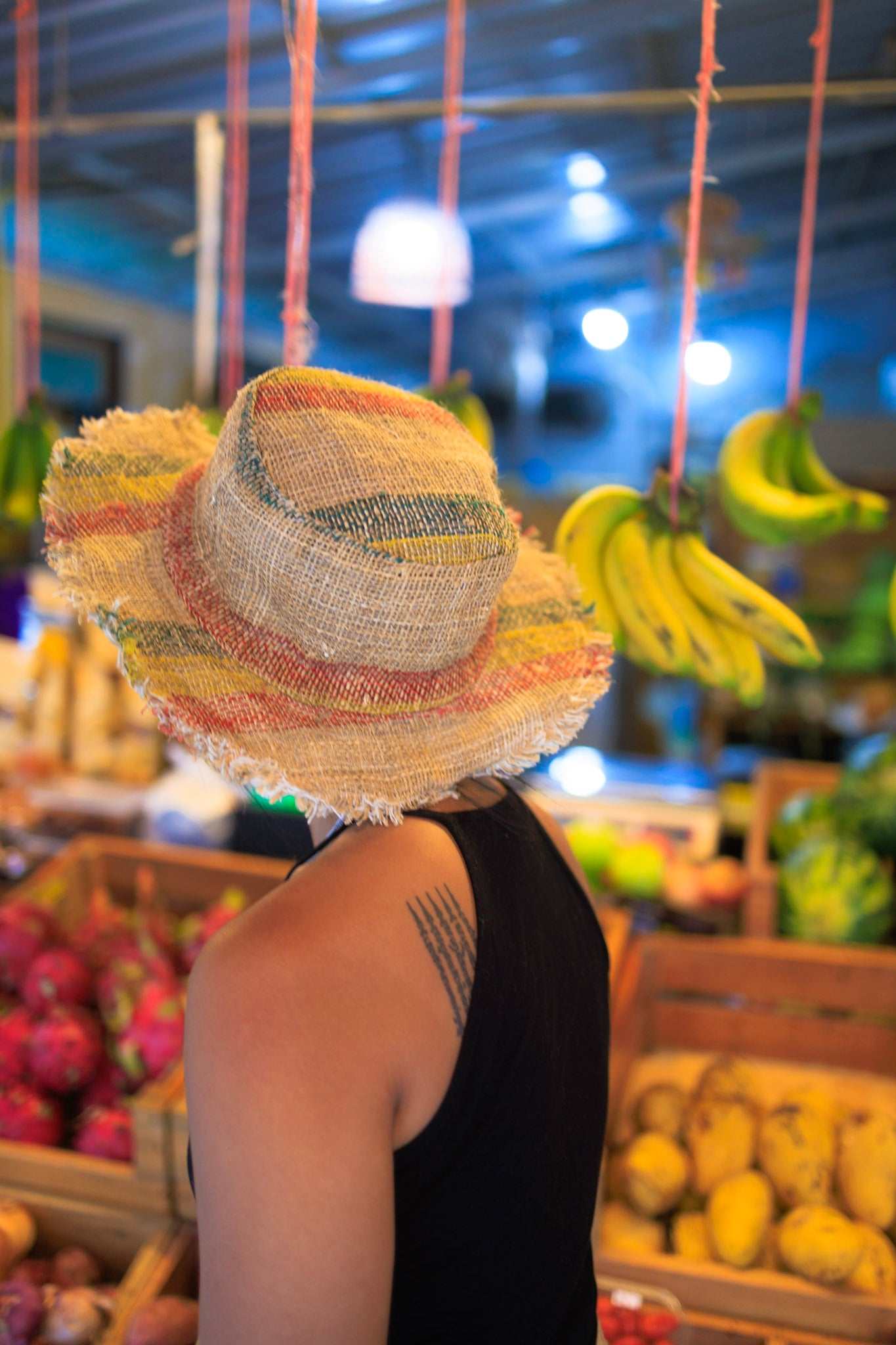 Person wearing a colorful straw hat in a market setting with fruits and vegetables.