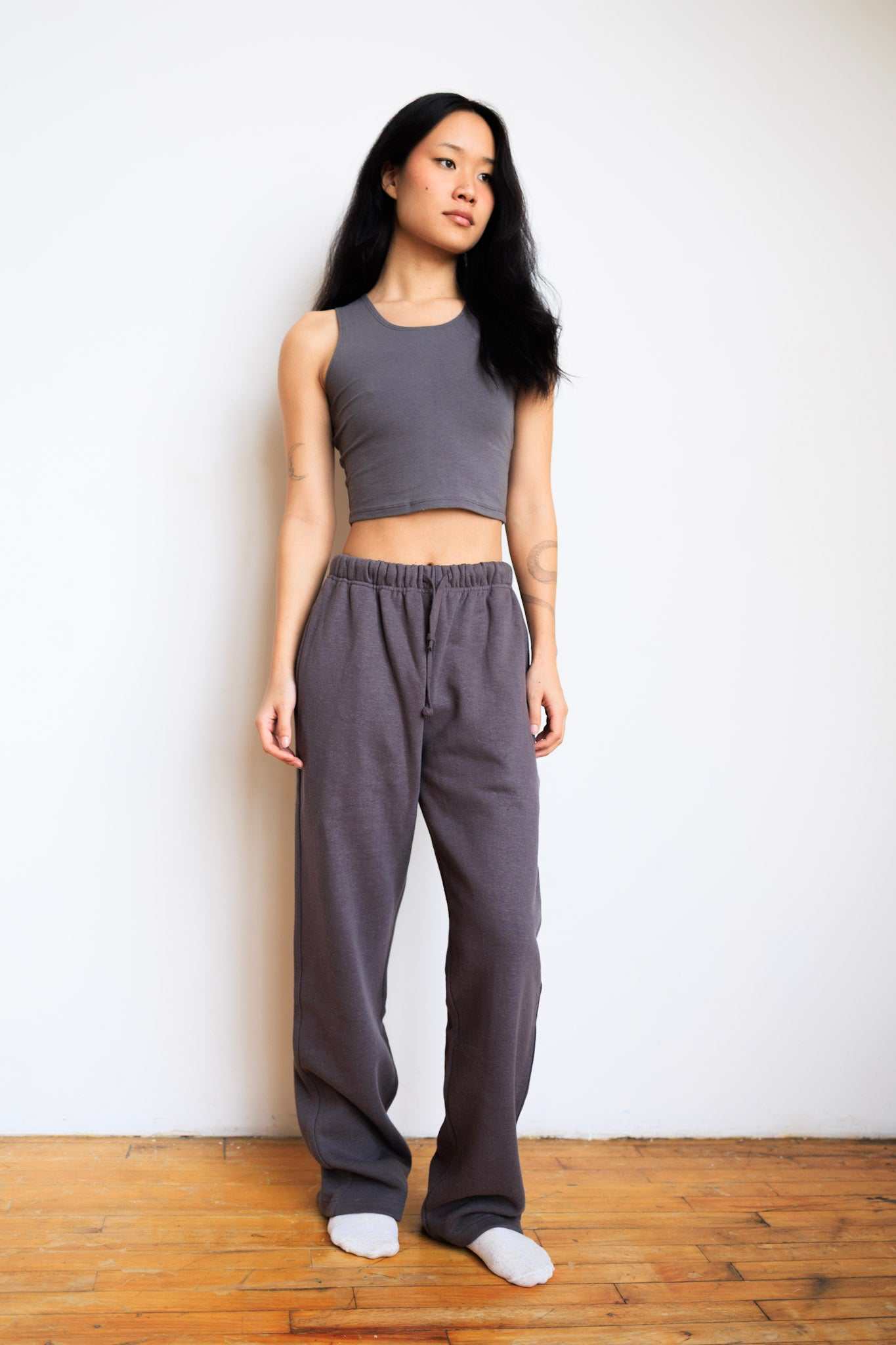 Woman wearing a gray tank top and pants standing against a white wall.