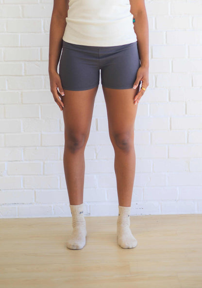 A person standing in front of a white brick wall wearing black active shorts with a visible logo on the waistband, paired with a white top and beige socks.