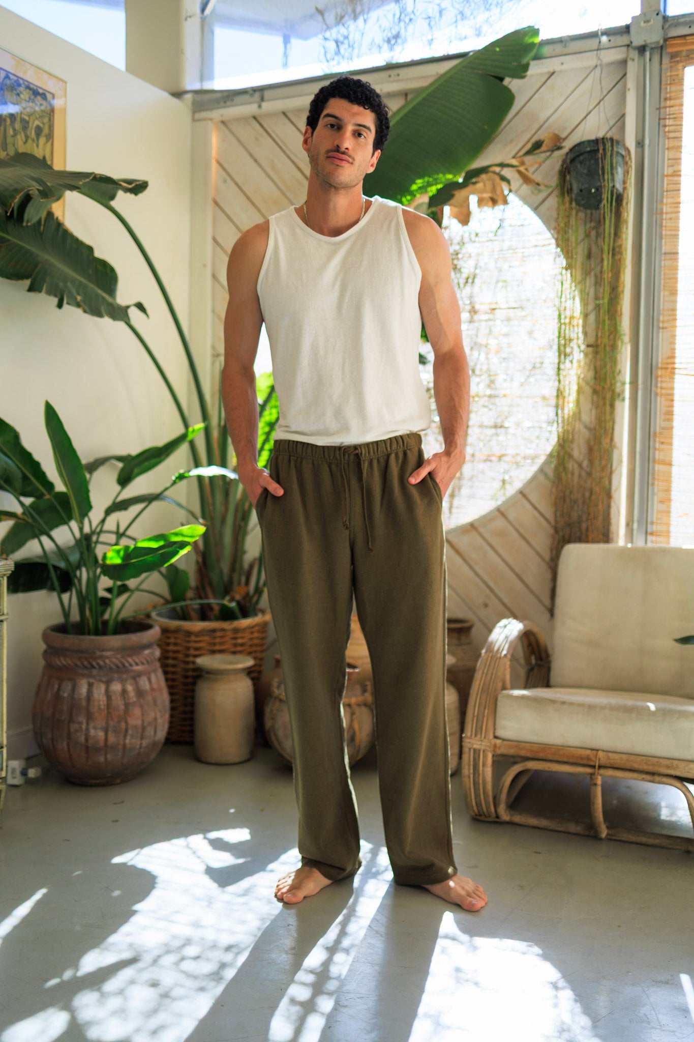 Man wearing olive hemp pants and white tank top in stylish indoor setting with plants and natural light