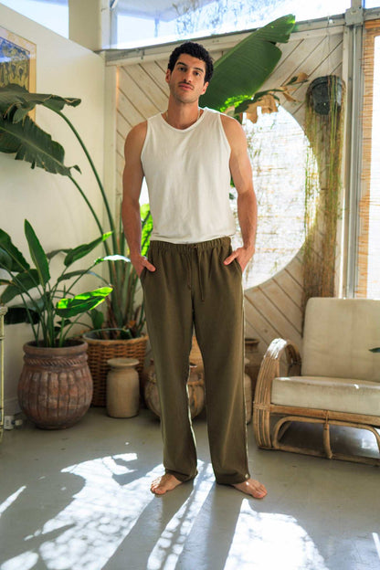 Man wearing olive hemp pants and white tank top in stylish indoor setting with plants and natural light